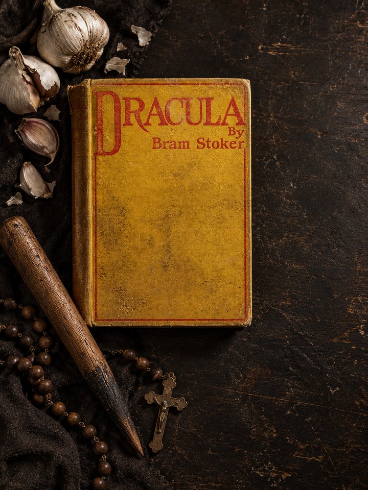 The yellow-cloth facsimile of the 1897 first edition of Dracula, its red ruled cover lettering reading Dracula by Bram Stoker, lying on a dark wooden surface beside bulbs of garlic, a wooden stake and a rosary with a crucifix.