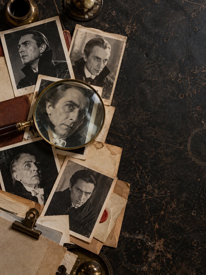 Old black-and-white portrait photographs of different screen Counts scattered across a dark desk, one magnified under a brass-rimmed magnifying glass, beside aged papers, a red wax seal and a bulldog clip.
